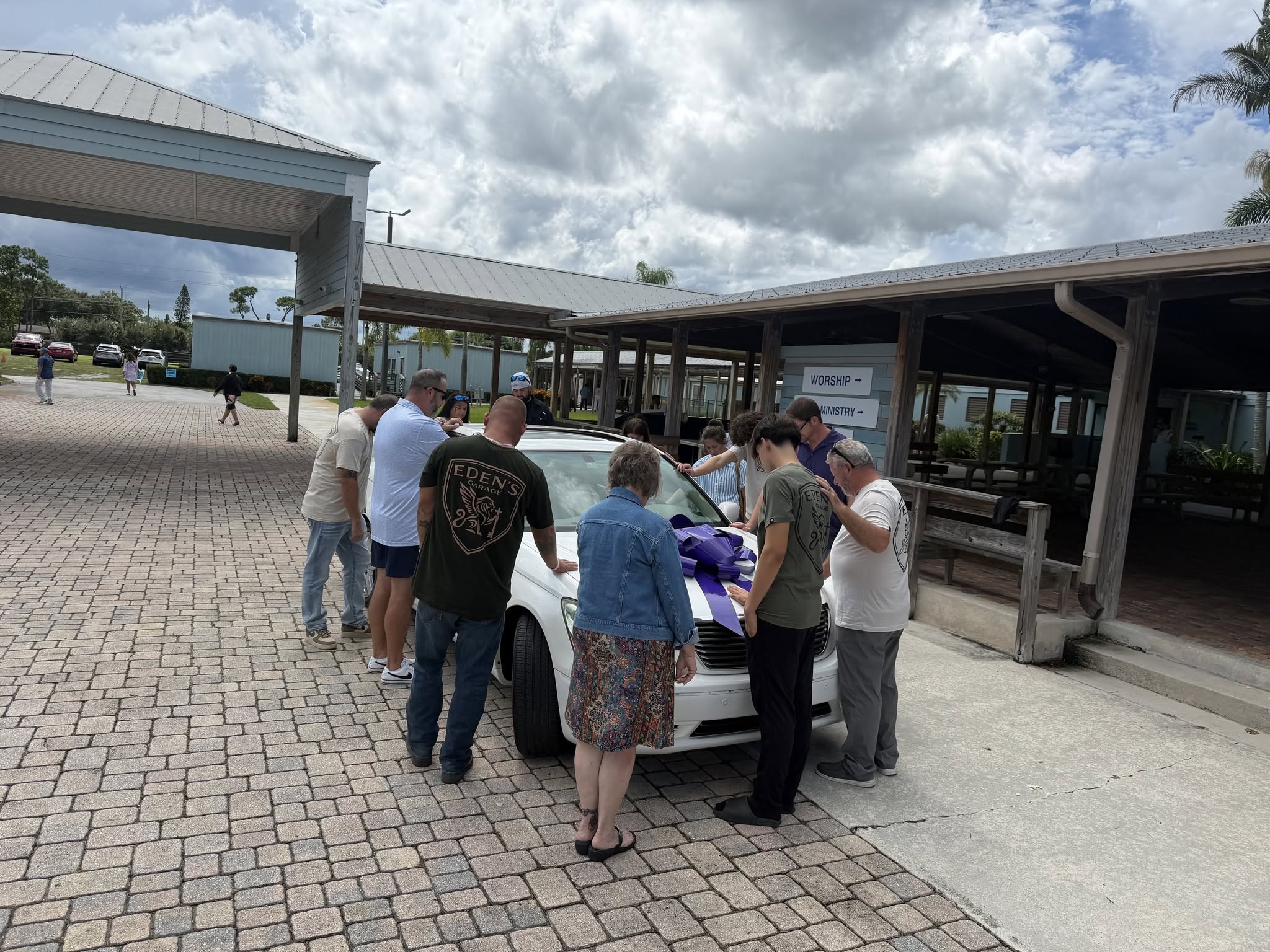 Family and volunteers rest their hands on a white car with a purple bow and pray over it before it is given away.